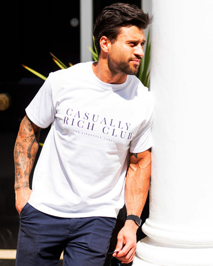 Man wearing a white t-shirt with 'Casually Rich Club' text, leaning against a white column.