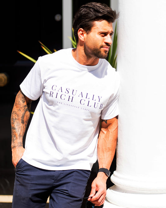 Man wearing a white t-shirt with 'Casually Rich Club' text, leaning against a white column.